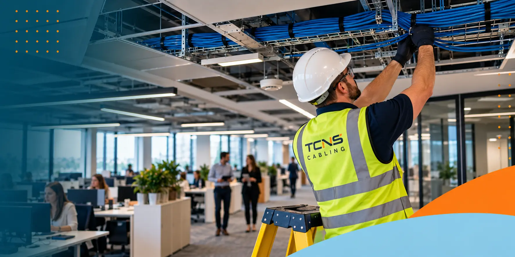 Engineer installing structured cabling in a live occupied office, standing on a ladder and working on overhead blue network cables while staff continue working in the background.
